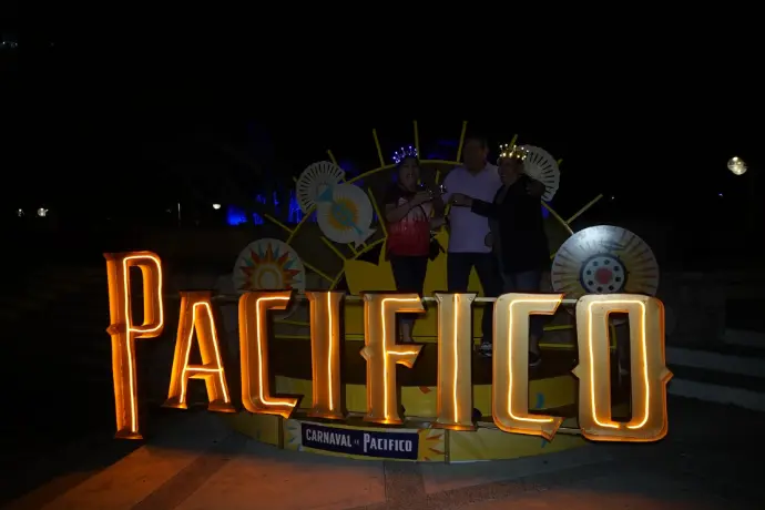 Photo Opp iluminado durante la noche, con una familia en la plaza disfrutando del Carnaval de Mazatlán 2026.