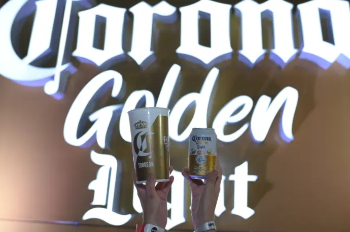 Vaso y lata de Corona Golden Light alzados frente al letrero luminoso de la marca en la Feria de Torreón 2024.