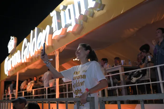 Asistente tomando selfie desde la Terraza VIP de Corona Golden Light con el letrero iluminado de la marca al fondo, Feria de Torreón 2024.