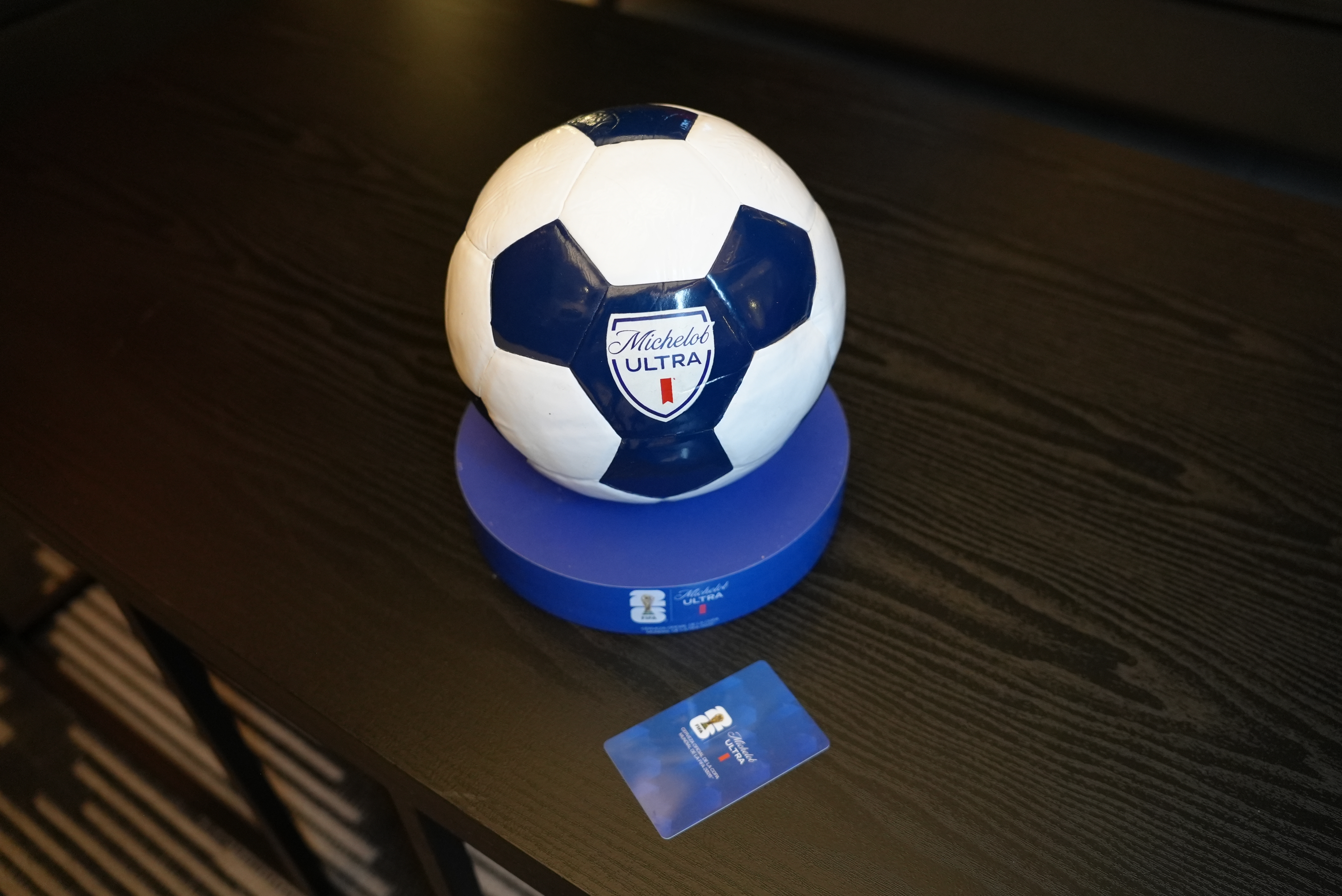 Balón Michelob Ultra y tarjeta llave en buró Balón de fútbol con logo Michelob Ultra sobre base circular junto a tarjeta llave FIFA World Cup 26 en buró de habitación de hotel