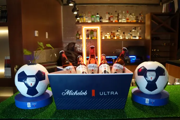 Balones y botellas Michelob Ultra en bar Balones de fútbol y botellas Michelob Ultra sobre pasto sintético junto a arco de neón en bar de hotel