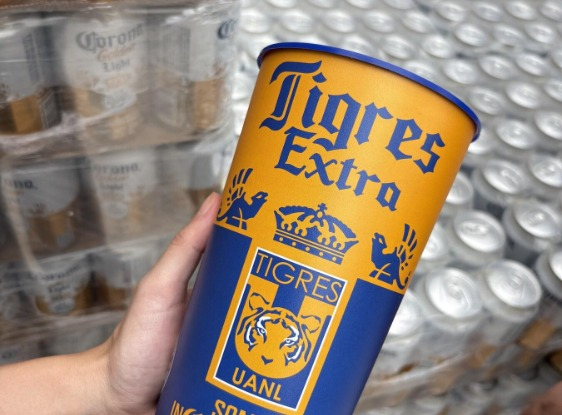 Vaso coleccionable Tigres Extra — Beer Experience 2026 Vaso coleccionable Tigres Extra en azul y oro con el escudo de Tigres UANL, sostenido frente al inventario de cerveza previo al inicio del partido.