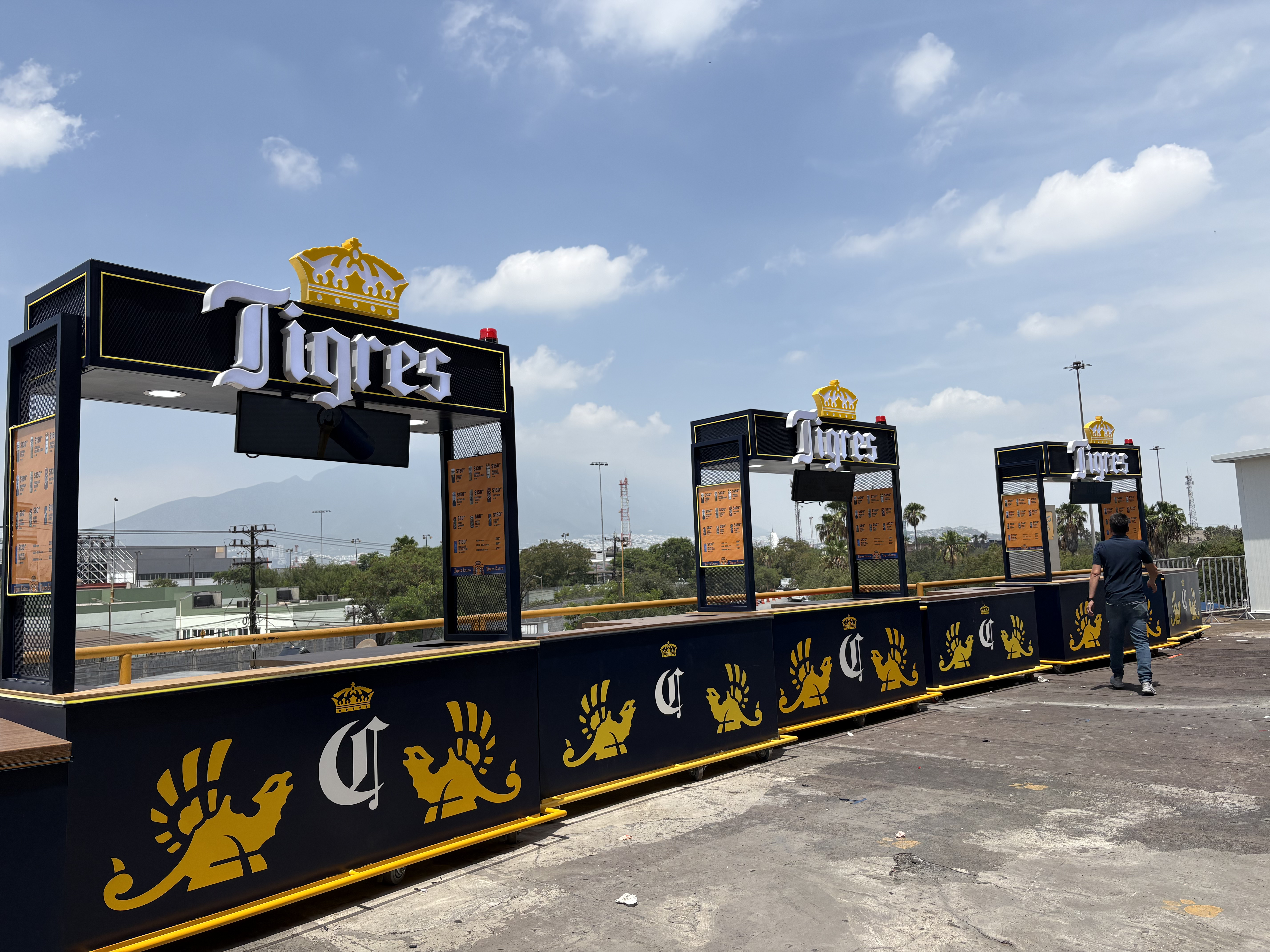 Fila de carritos móviles de punto de venta con letreros luminosos Tigres y branding Corona instalados en la explanada del Estadio Universitario, con el Cerro de la Silla al fondo.